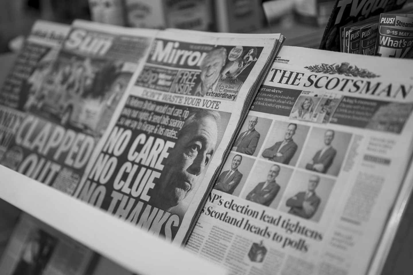Black and white photograph of newspaper front pages on a newsstand, showing the Mirror with a large image of Nigel Farage and the headline 'No Care No Clue No Thanks' alongside The Scotsman with election coverage.